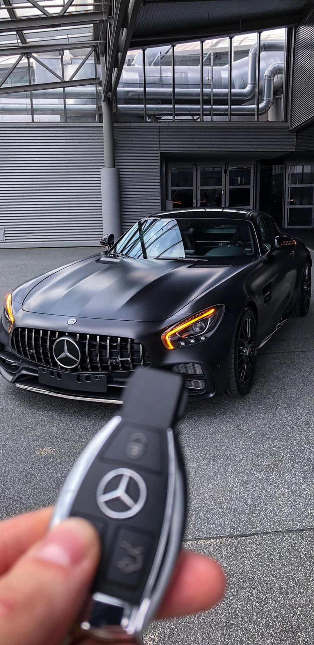 Mercedes-AMG GT key fob held in front of a matte black luxury sports car parked near a modern glass building facade.