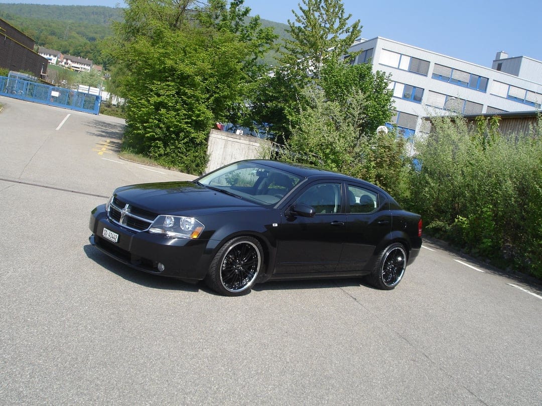 Black Dodge Avenger sedan with custom rims parked on an asphalt lot under a blue sky and green trees.