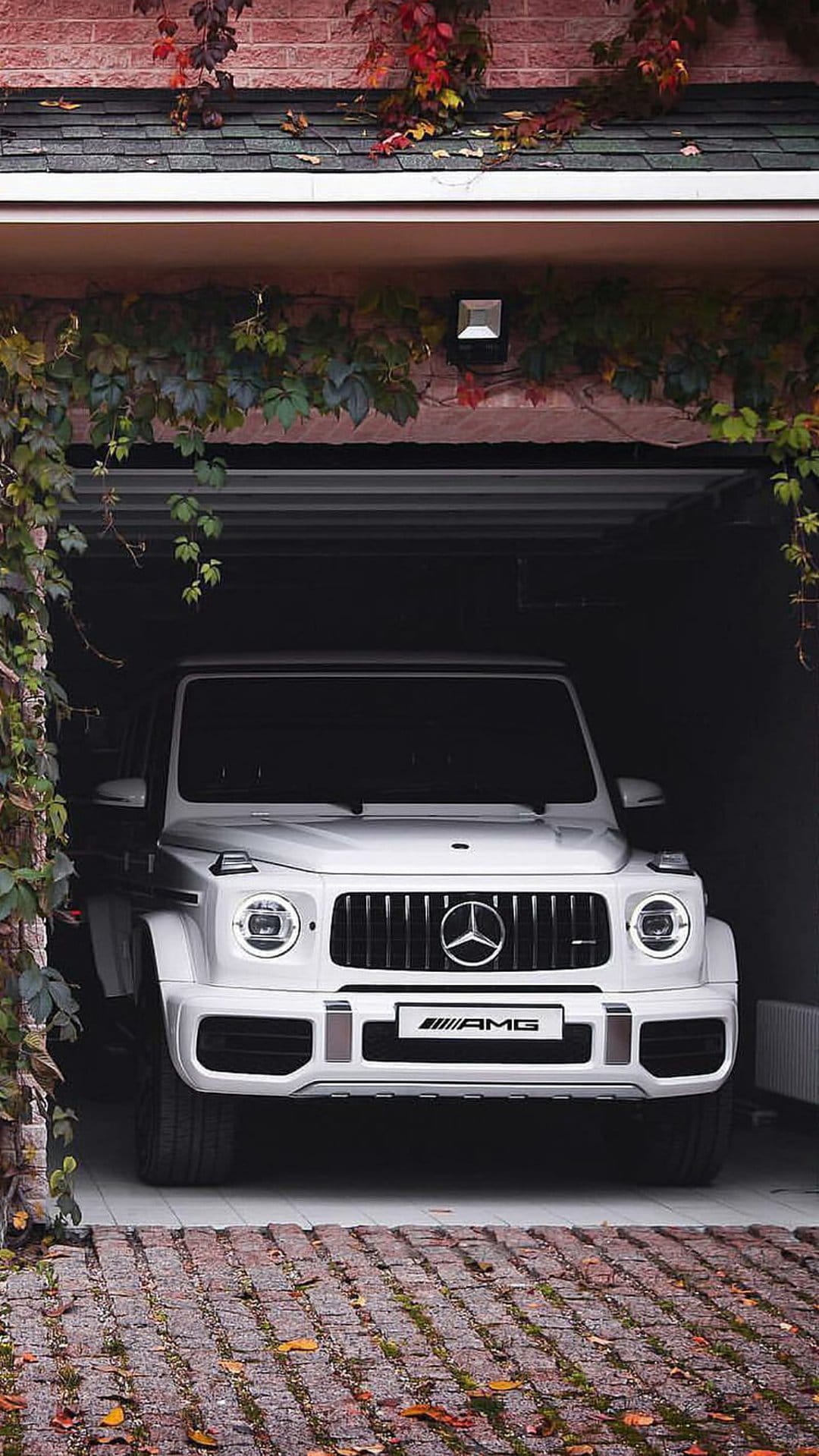 White Mercedes-AMG G 63 SUV parked in a dark garage framed by red and green autumn leaves and brick walls.