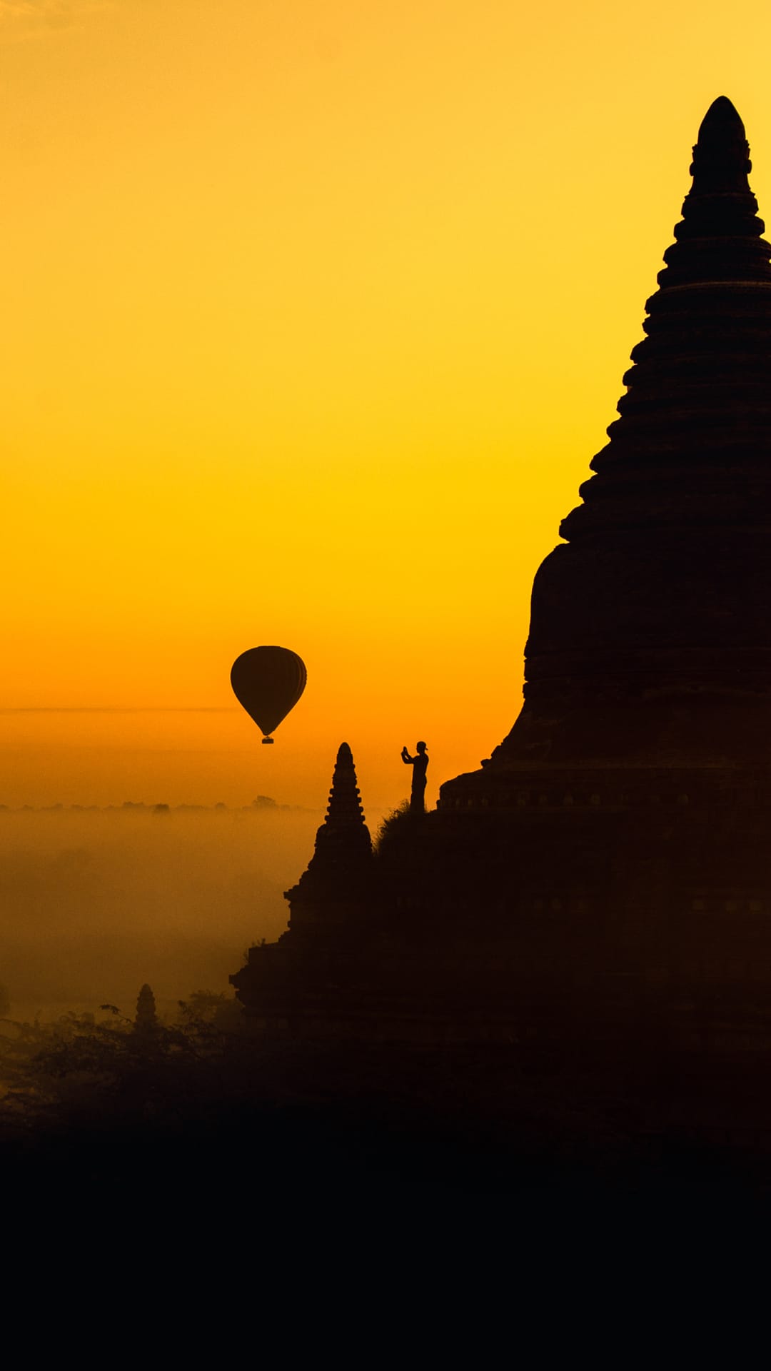 Ancient stupa silhouette and a hot air balloon against a golden sunrise sky with a person standing nearby.