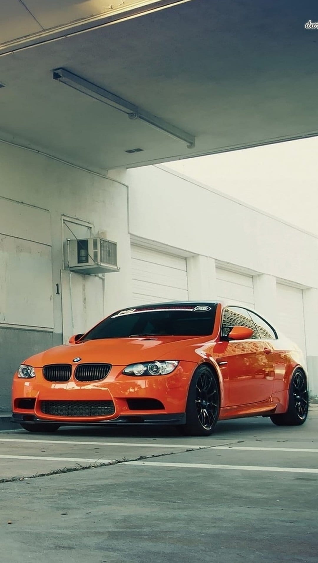 Orange BMW M3 Coupe parked in a modern industrial garage with fluorescent lights and black multi-spoke rims.