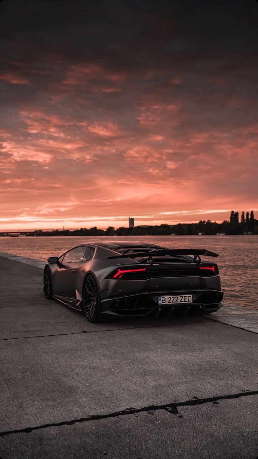 Lamborghini Huracan on a concrete pier at dusk with red taillights glowing against an orange sunset sky.