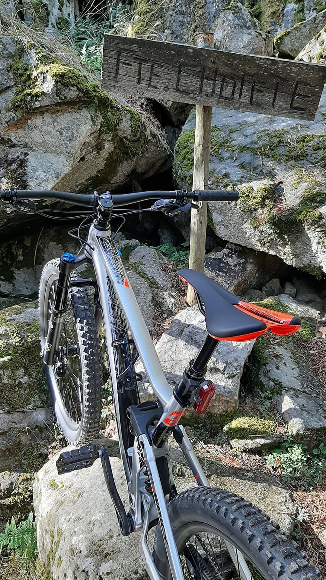 Silver mountain bike with orange accents parked beside a weathered wooden sign on a mossy rocky trail.