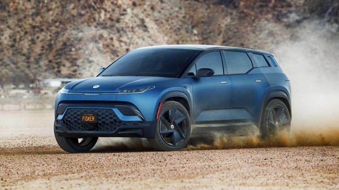 Blue Fisker Ocean SUV driving on a dirt road kicking up a cloud of dust against a rocky desert background.