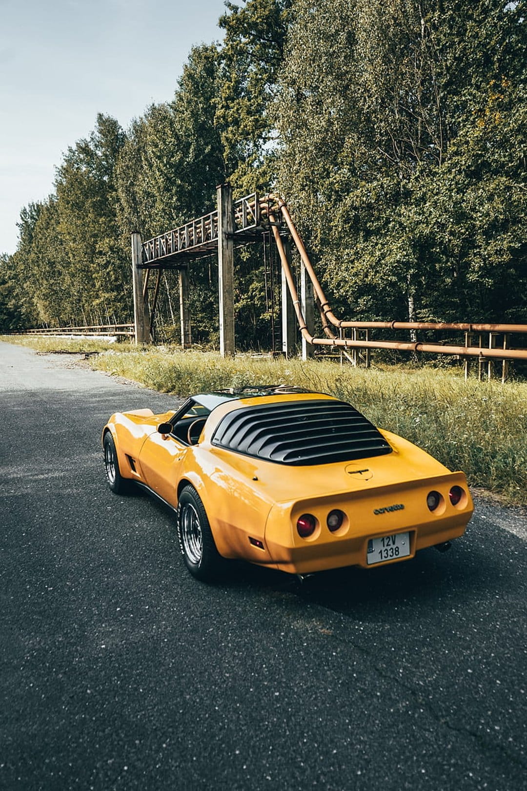 Yellow Chevrolet Corvette Stingray parked on an overgrown road near rusted industrial pipes and green trees.