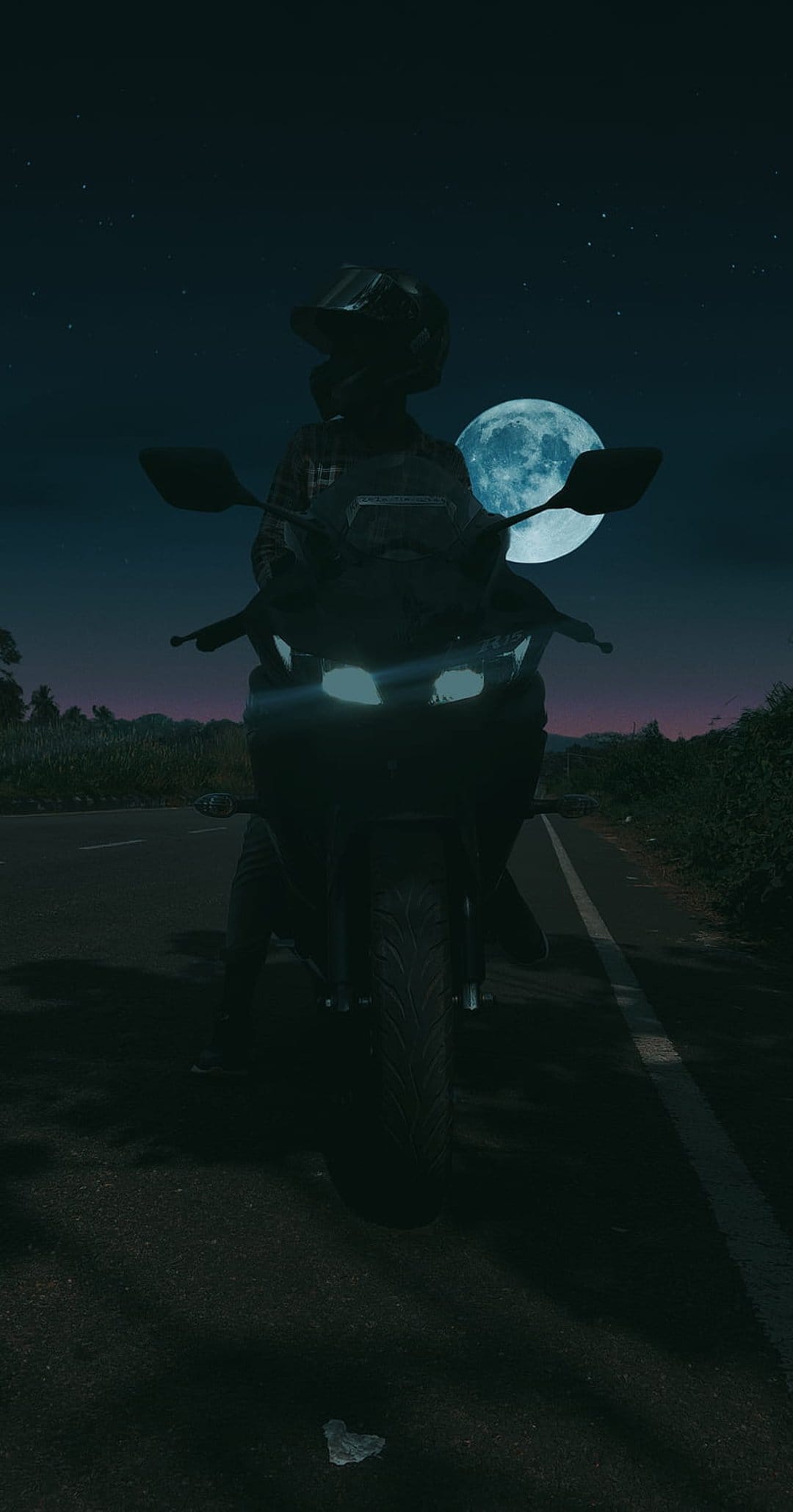 Motorcyclist in silhouette travels along a dark road toward a massive, bright full moon in a starry night sky.