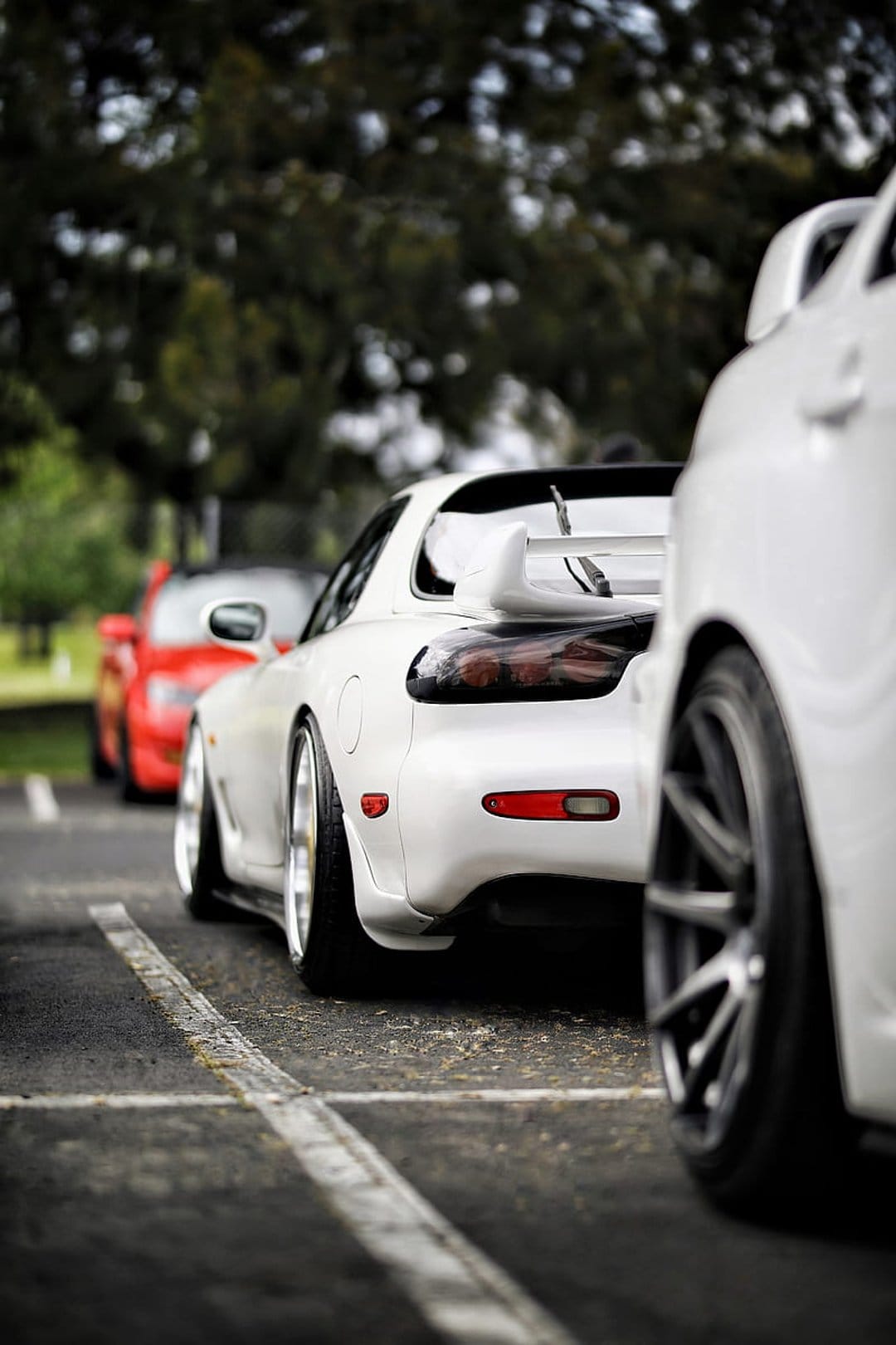 White Mazda RX-7 with a large rear wing parked on wet asphalt, featuring a blurred red car in the background.