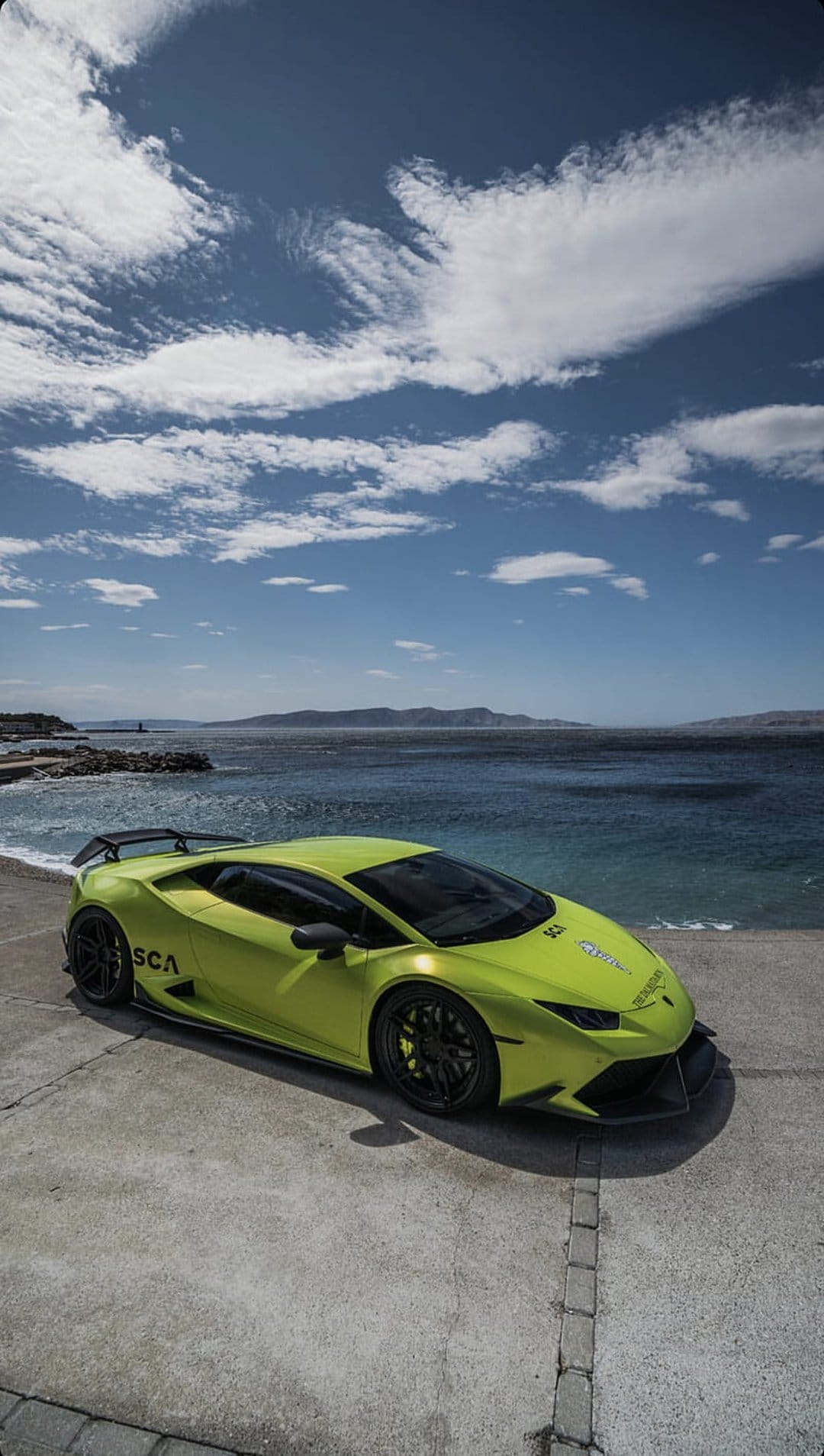 Lime green Lamborghini Huracan with black wheels parked on a concrete overlook against a blue ocean and sky.
