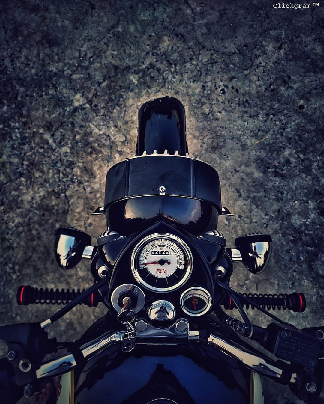 Chrome motorcycle handlebars and an analog speedometer sit over a dark, textured stone background in a top view.