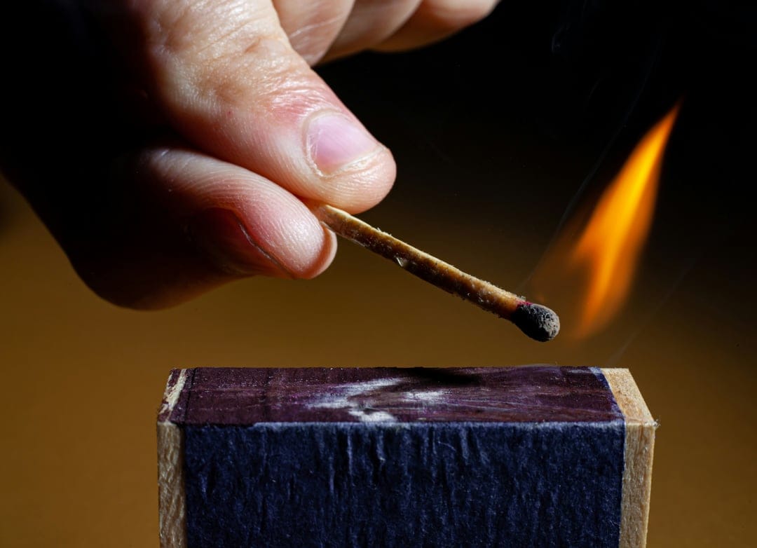 Hand striking a matchstick against a matchbox creates a bright orange flame and thin smoke on a dark backdrop.