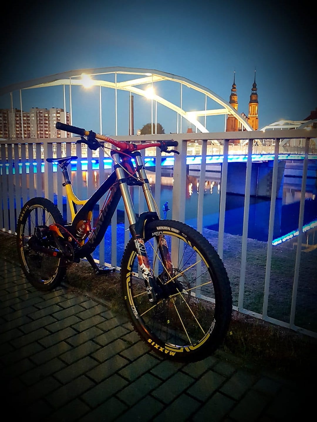 Downhill mountain bike rests on cobblestones near an illuminated blue bridge under a dusky urban sky.