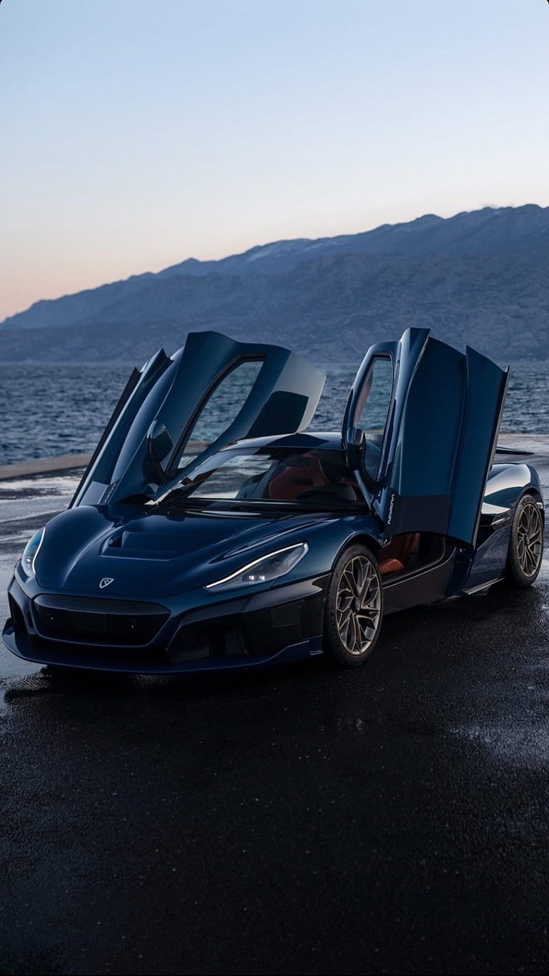 Blue Rimac Nevera hypercar with open butterfly doors parked near a tranquil sea under a clear mountain horizon.