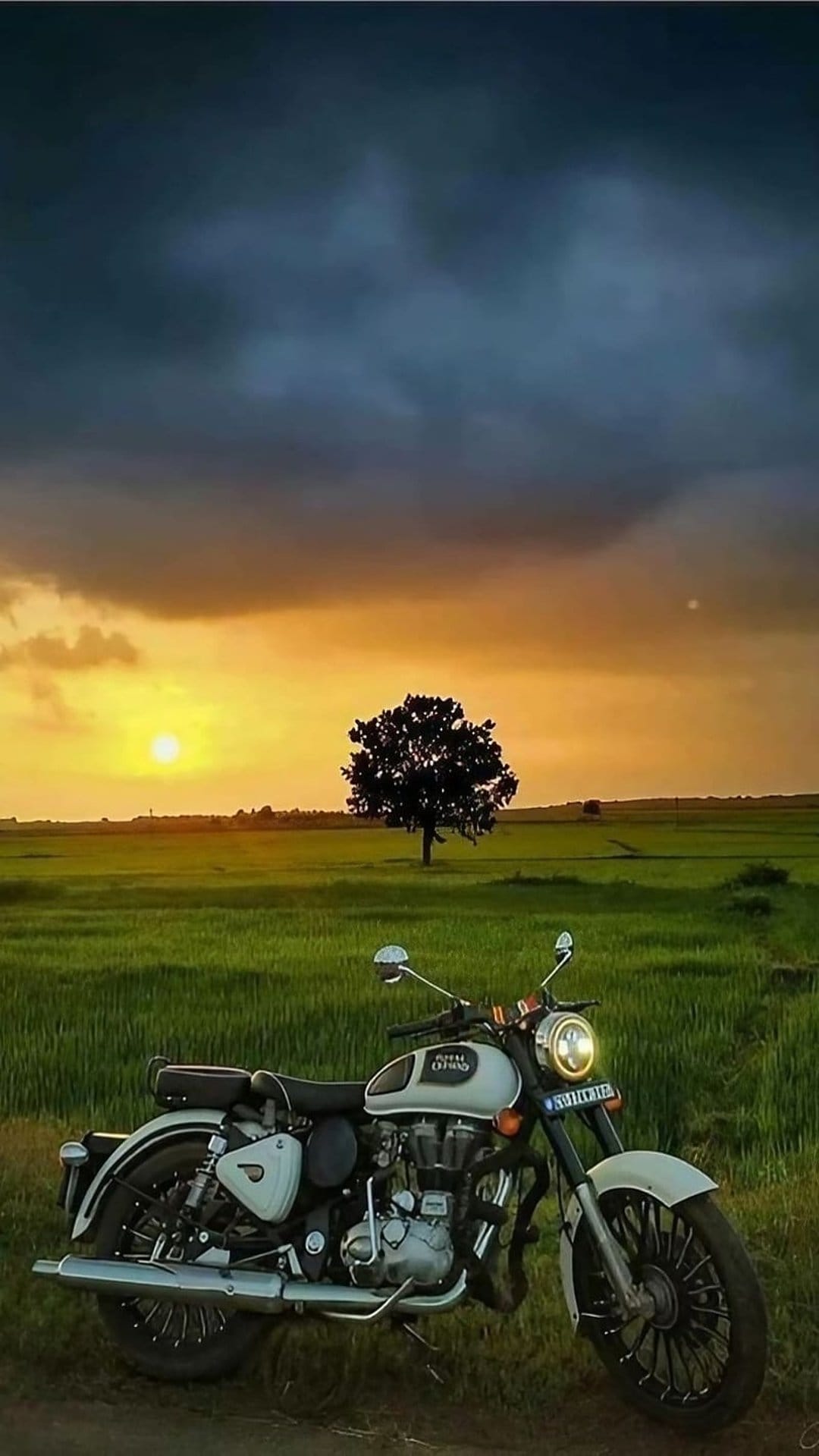 White motorcycle with its headlight on stands in a grassy field under a cloudy orange and yellow sunset sky.