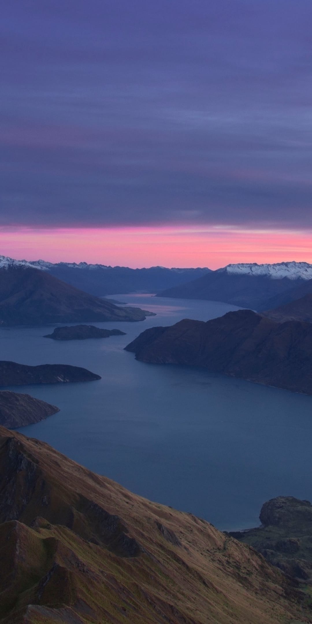 Rugged mountain ridges and a winding lake reflect a vibrant pink and purple twilight sky with snowy peaks.