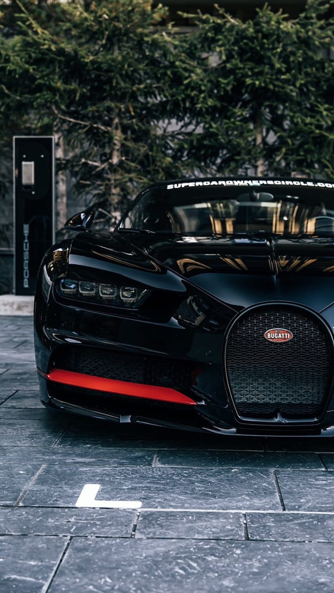 Black Bugatti Chiron with a red front accent parked on stone pavement next to a white charging station and greenery.