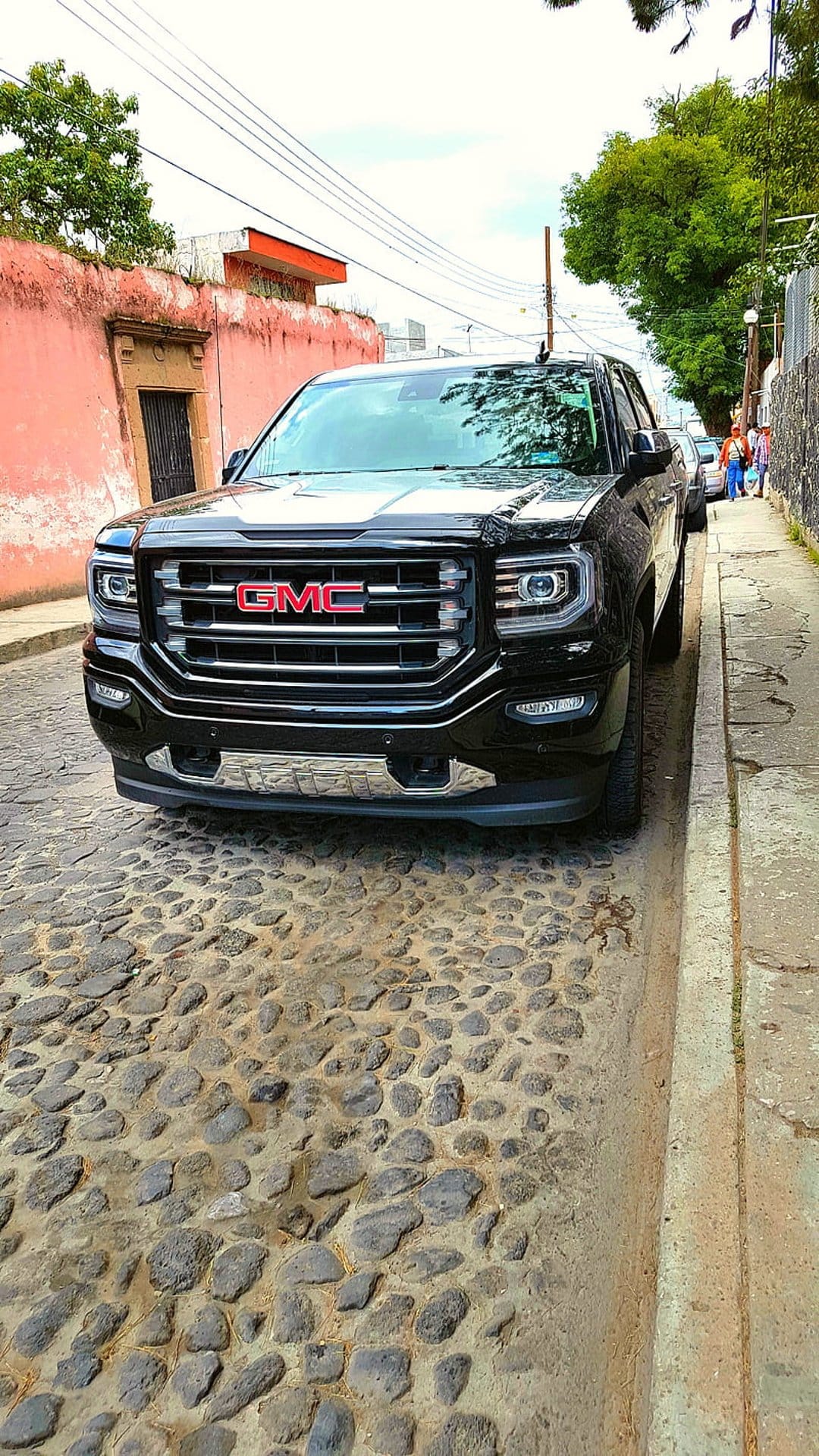 Black GMC Sierra pickup truck with a chrome grille parked on a cobblestone road in front of a pink building.