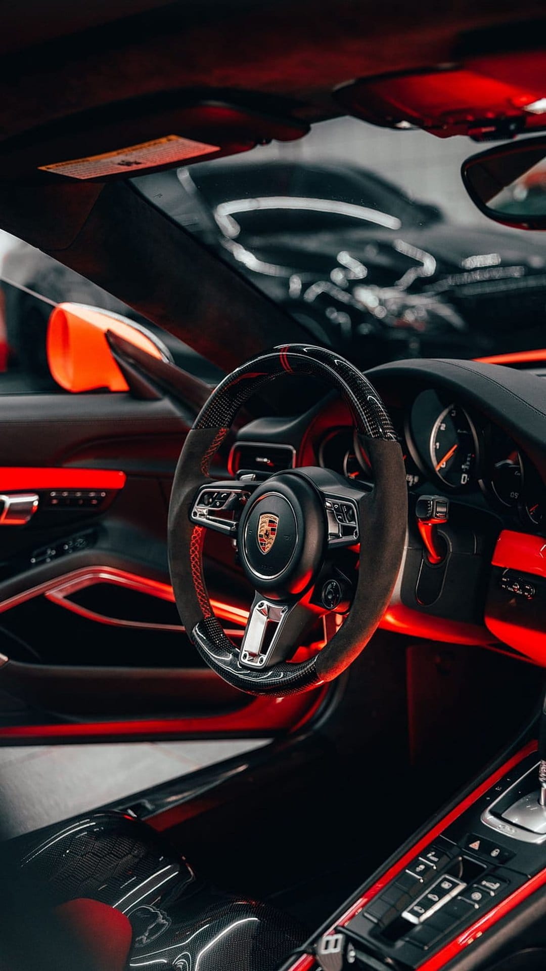 Porsche steering wheel with red stitching and emblem illuminated by scarlet ambient lights in a dark car cabin.