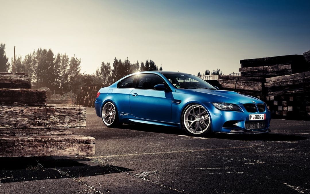 Blue BMW M3 E92 coupe parked between stacks of wood in low sunlight with metallic paint and silver wheels.
