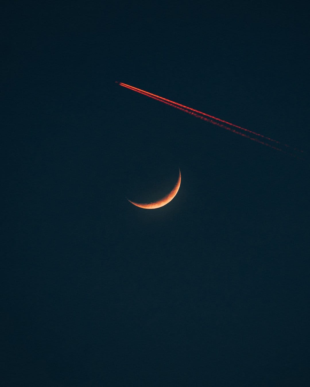 Slender orange crescent moon glows beneath a vivid red jet contrail streaking across a dark black night sky.