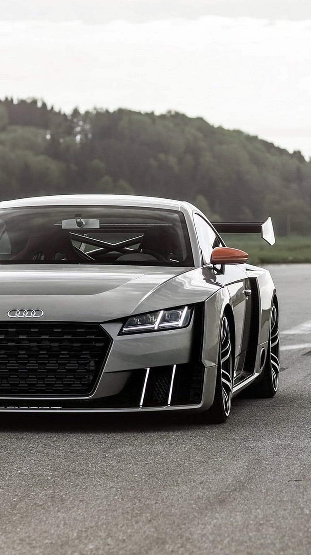 Audi TT RS-MR race car with a large rear wing, orange mirror caps, and a silver body parked on an asphalt track.