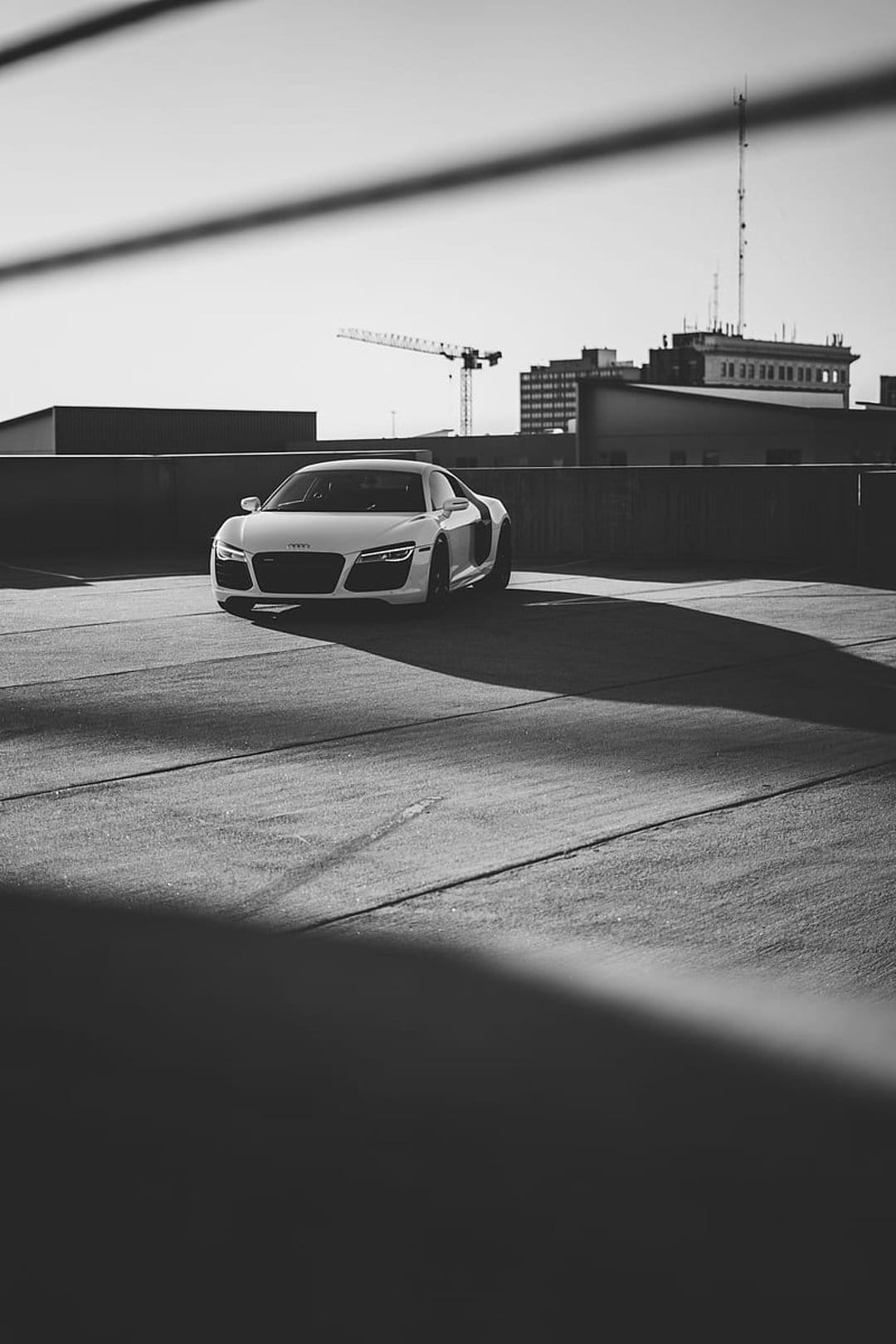 White Audi R8 sports car parked on a concrete rooftop with harsh shadows and a distant crane in monochrome.
