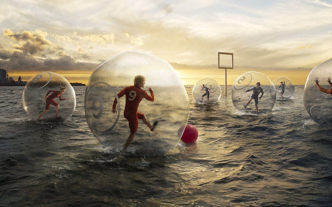 Runners inside transparent spheres compete on the ocean surface under a bright orange sunset with a distant city.