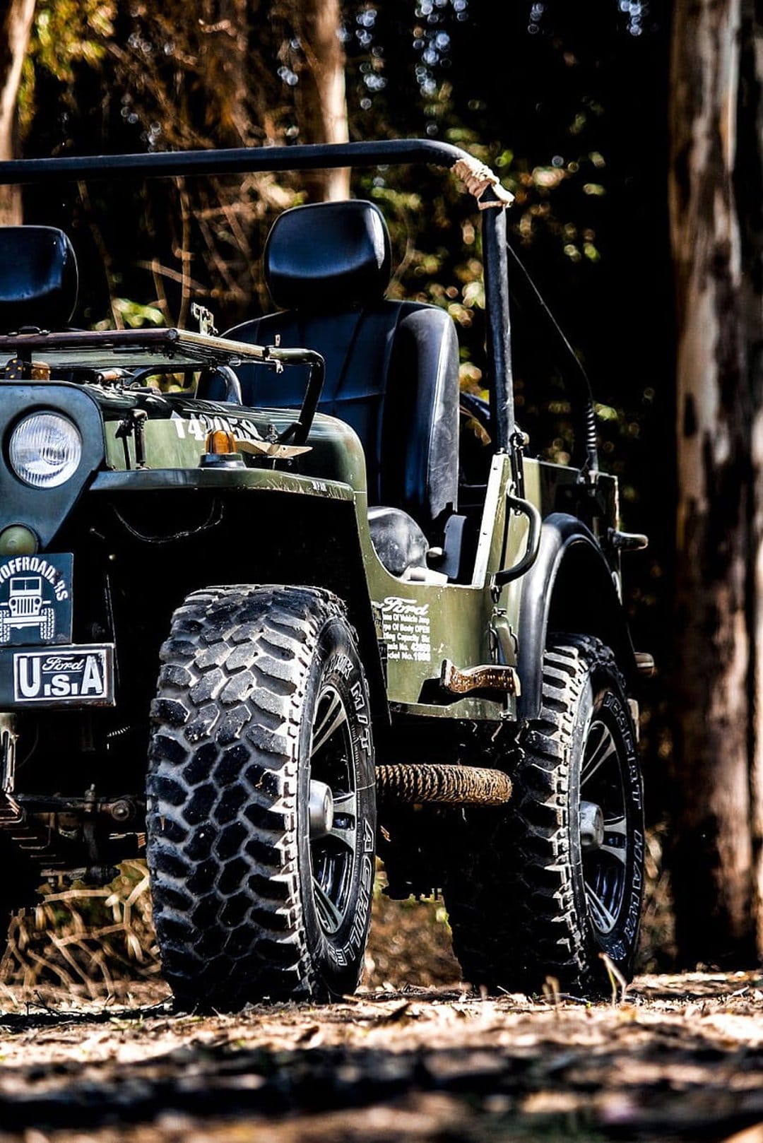 Vintage olive green Jeep with off-road tires parked on a dirt path under dappled sunlight in a lush forest.