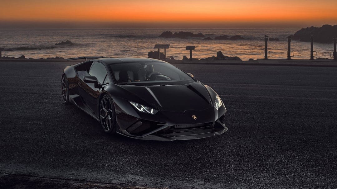 Black Lamborghini supercar on a coastal road at sunset with orange skies and ocean waves in the background.