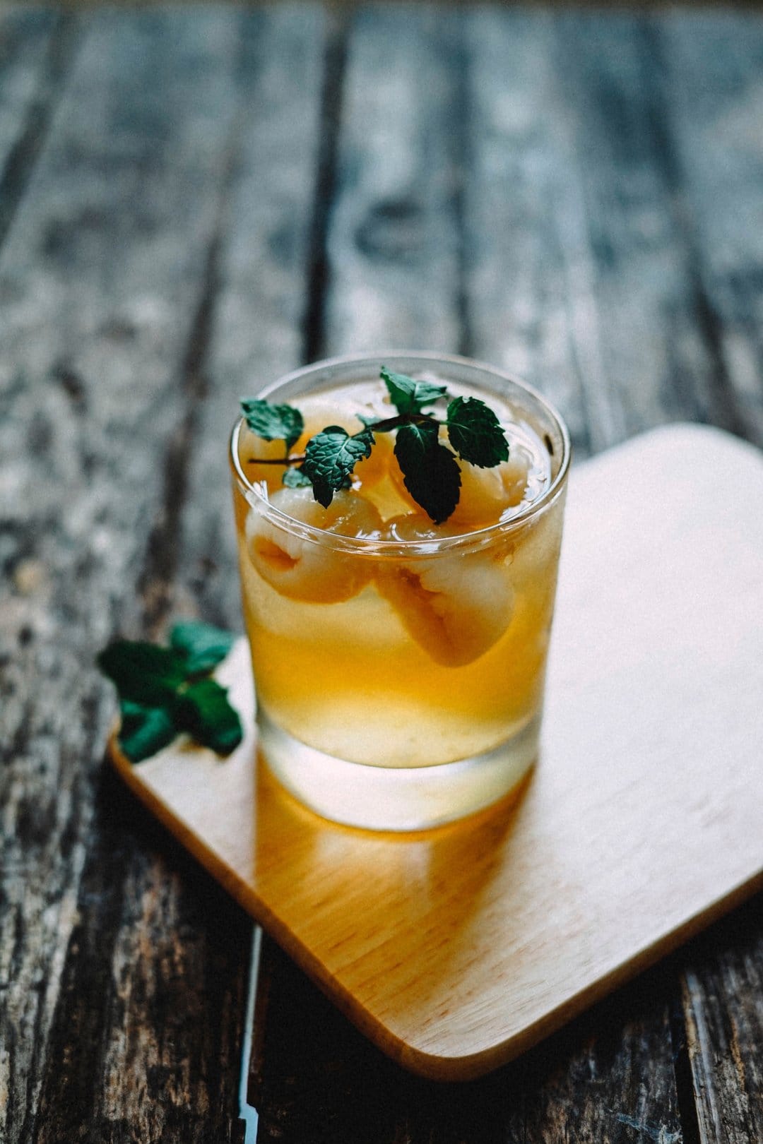 Clear glass of golden lychee juice with mint leaves and fruit on a wooden coaster atop a dark rustic table surface.