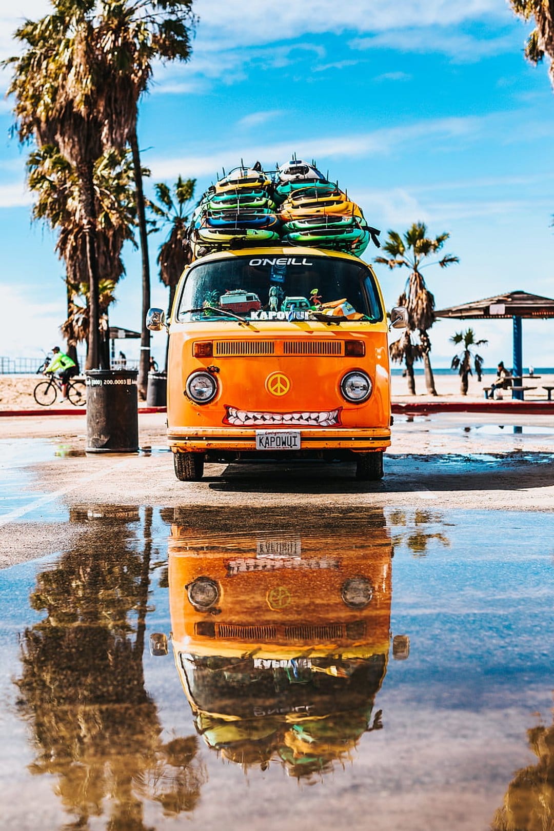 Orange vintage VW bus with surfboards reflects in a clear puddle on a sunny coastal road near the ocean.
