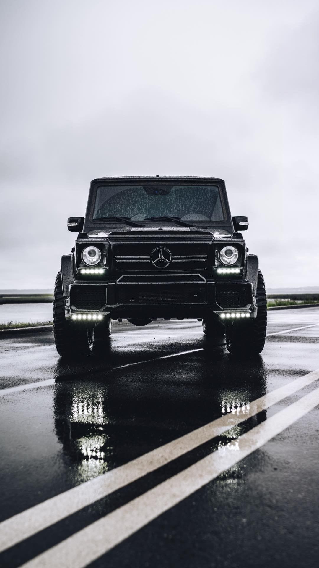 Front view of a black Mercedes-Benz G-Class with glowing LED lights reflecting on a glistening wet rainy road.