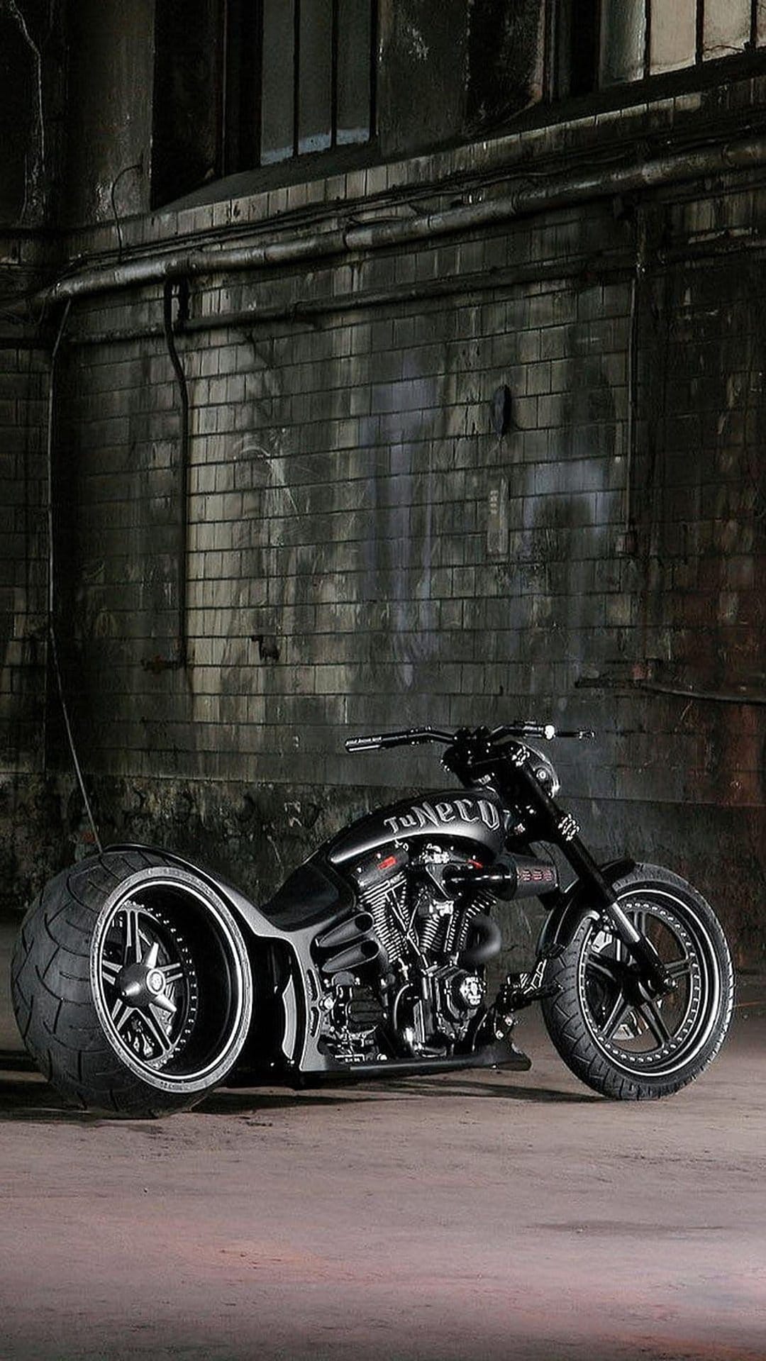 Black custom cruiser motorcycle with a wide rear tire parked inside a dimly lit industrial brick warehouse.