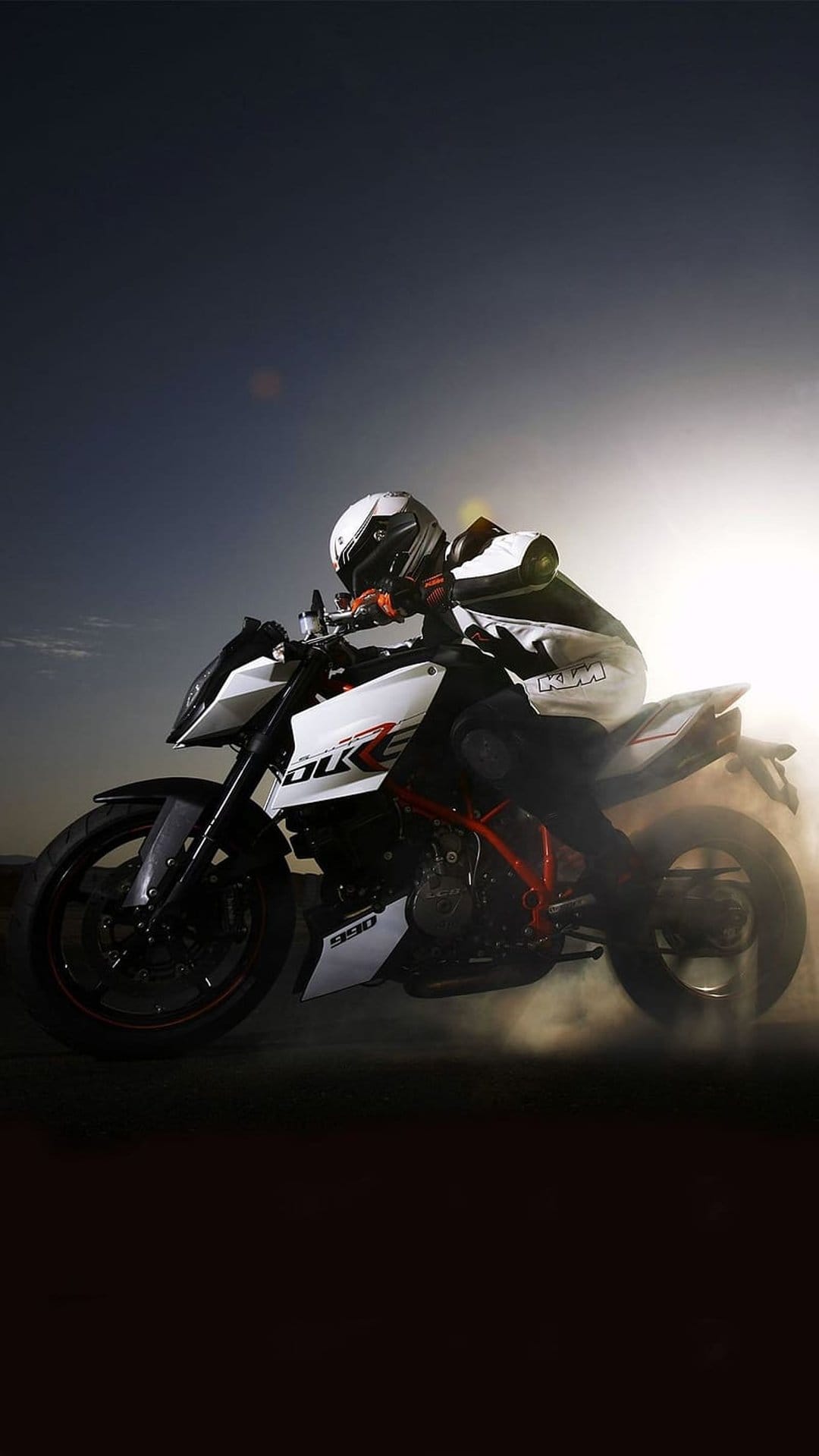 Motorcycle rider on a KTM Duke leans into a sharp turn as dust and smoke billow under a bright sunlit sky.