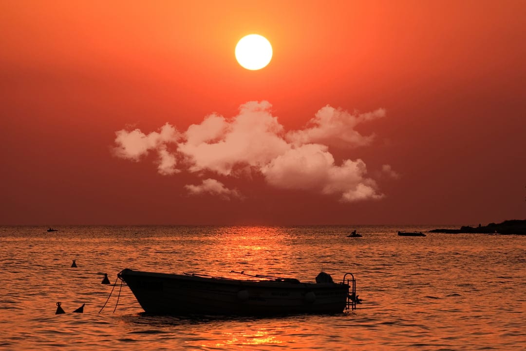 Lone boat silhouetted against a fiery orange and red sunset over calm ocean waters with wispy clouds.