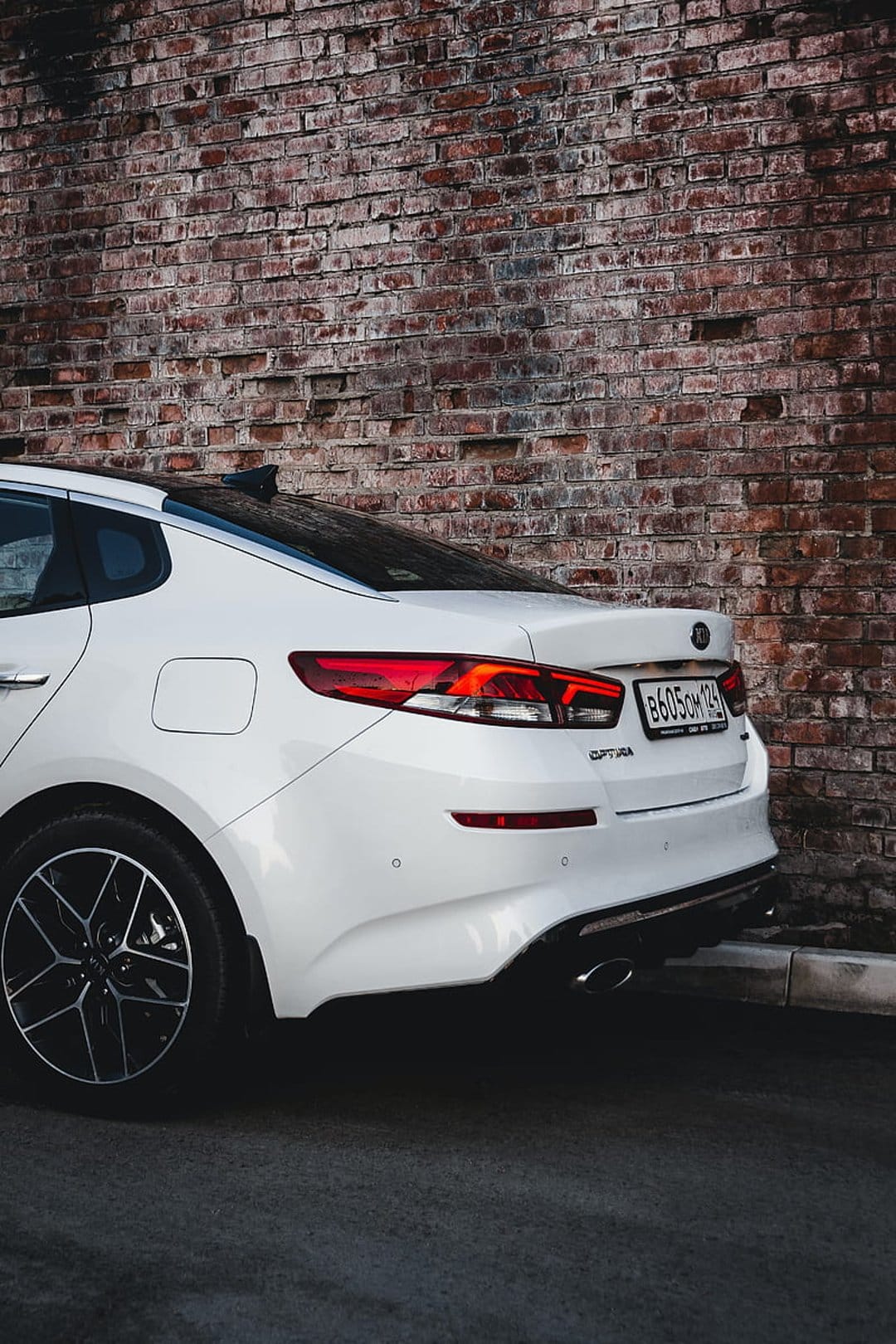 White Kia Optima rear profile with red taillights parked on asphalt beside a textured weathered brick wall.