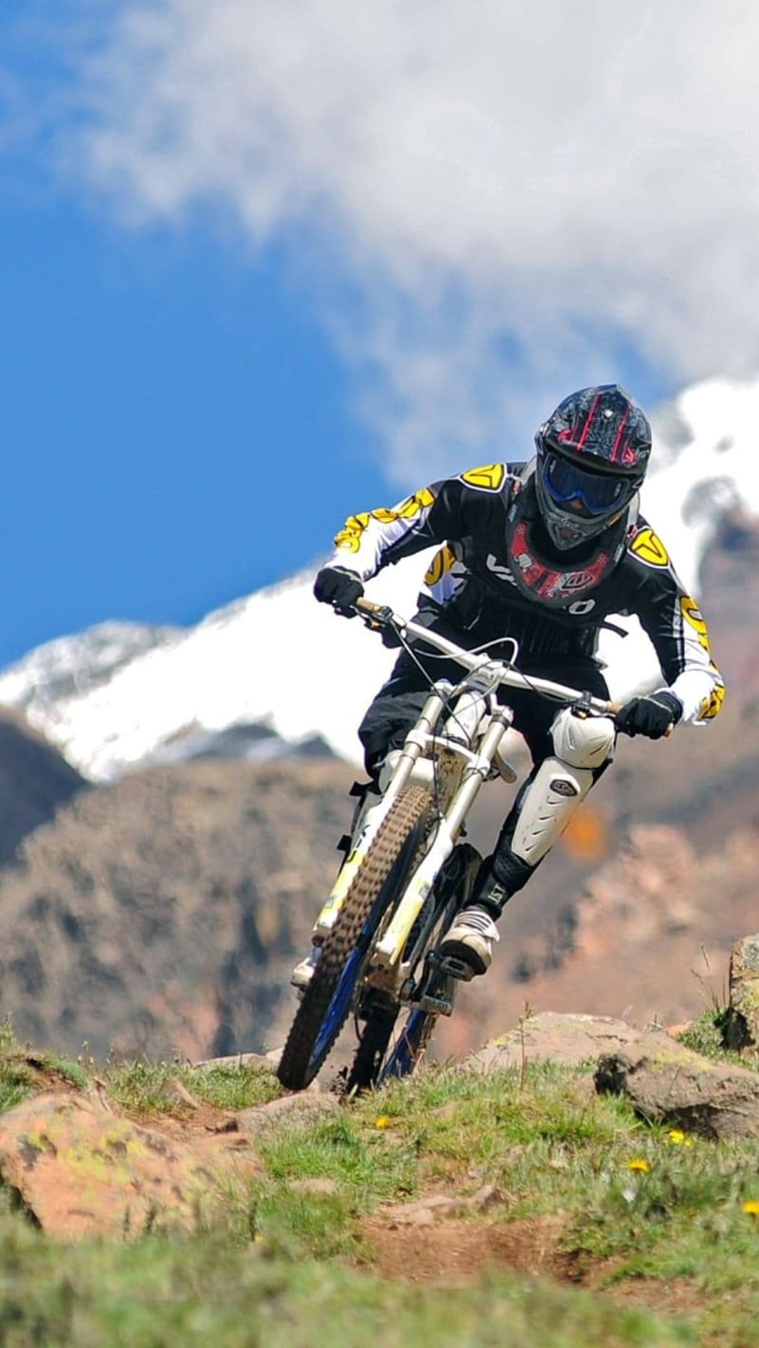 Mountain biker in full gear descends a rocky mountain ridge under a blue sky with snowy peaks in the distance.