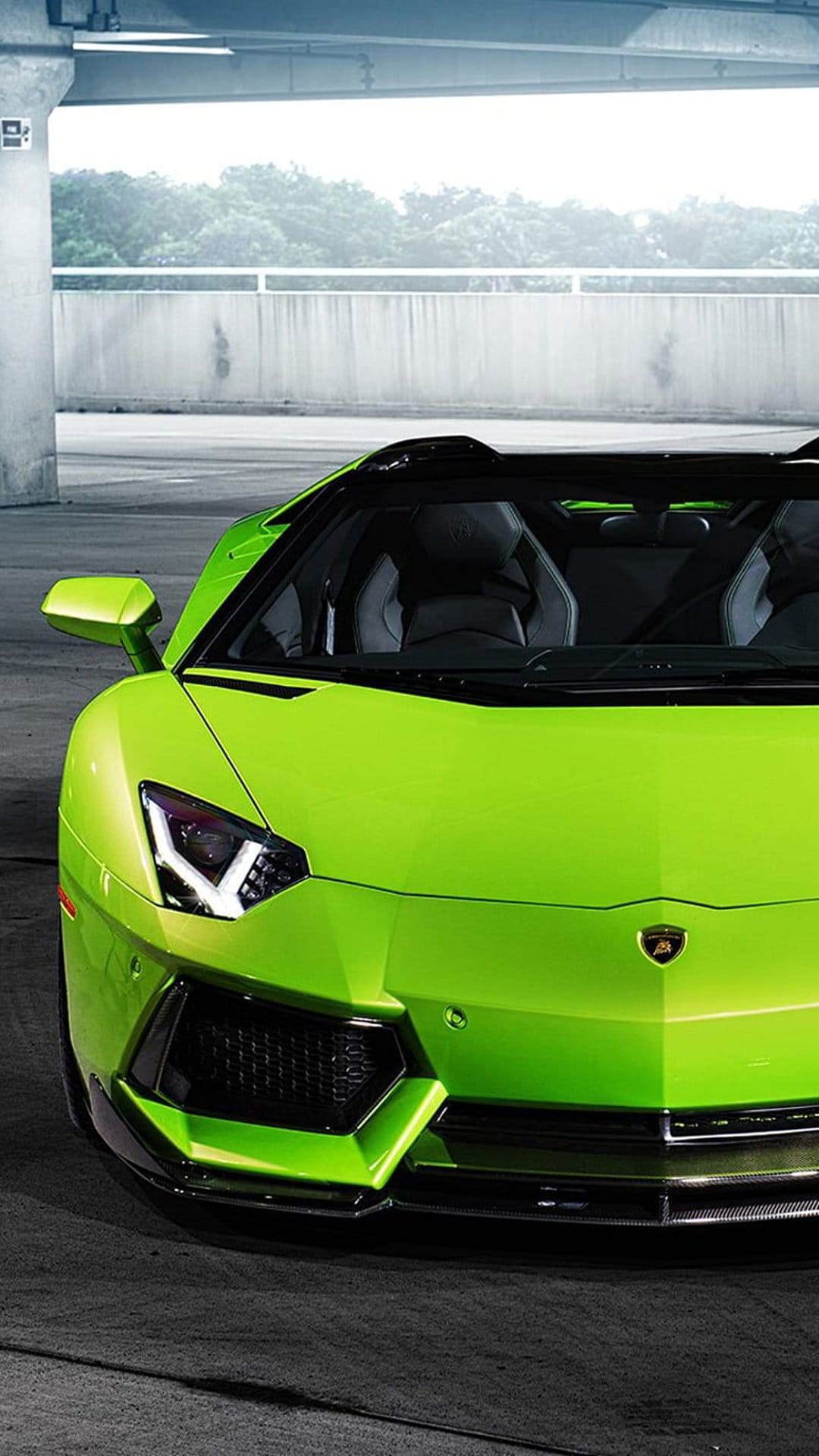 Lime green Lamborghini Aventador Roadster with glowing headlights parked near a concrete wall and greenery.