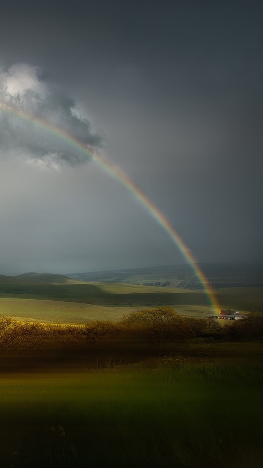 Colorful rainbow arches over a stormy sky and green rolling hills with a small white farmhouse in the distance.