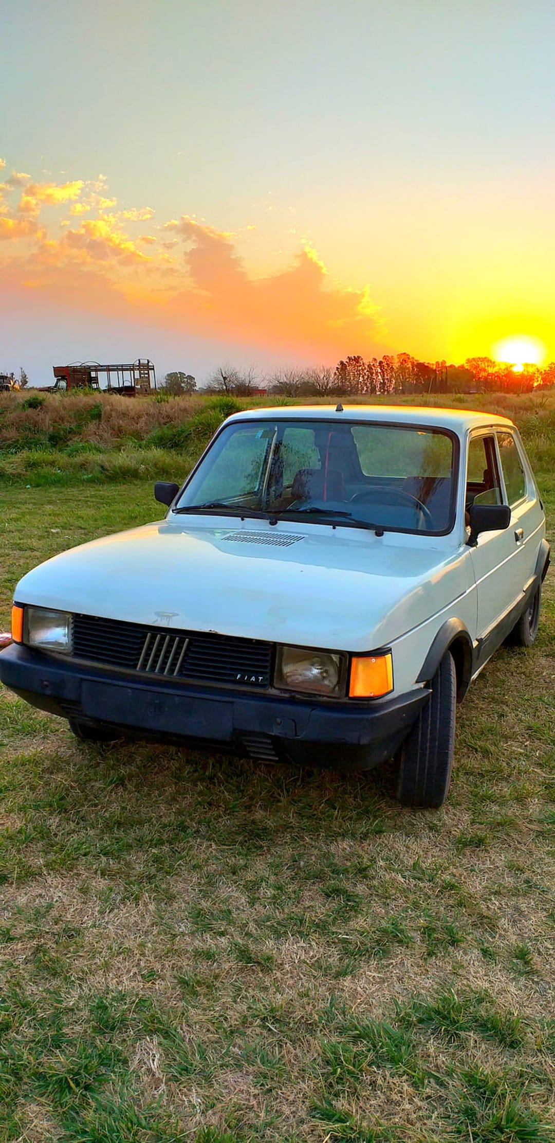 White vintage Fiat car parked in tall grass against a vibrant orange and yellow sunset sky during golden hour.