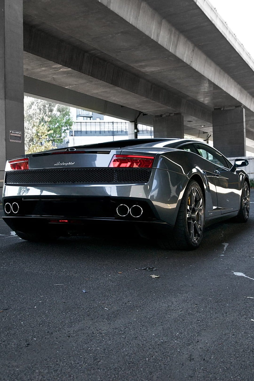 Dark grey Lamborghini rear view under a concrete overpass with glowing red taillights and dual exhausts.