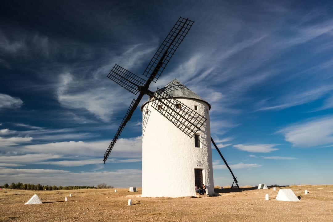 Traditional white windmill with dark wooden sails under a blue sky with thin clouds in a Spanish landscape.