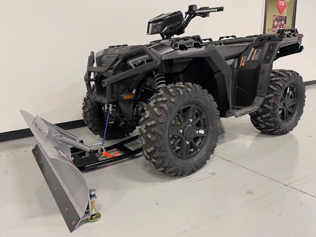 Matte black Polaris ATV with a silver snow plow blade and aggressive tires in a bright indoor showroom.