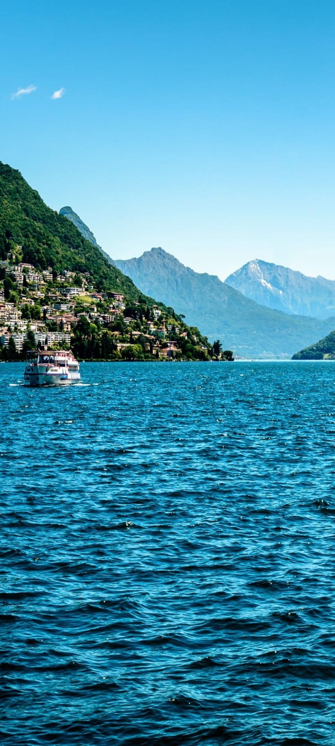 White pleasure boat sails past colorful houses on a steep mountainside overlooking the blue waters of Lake Como.