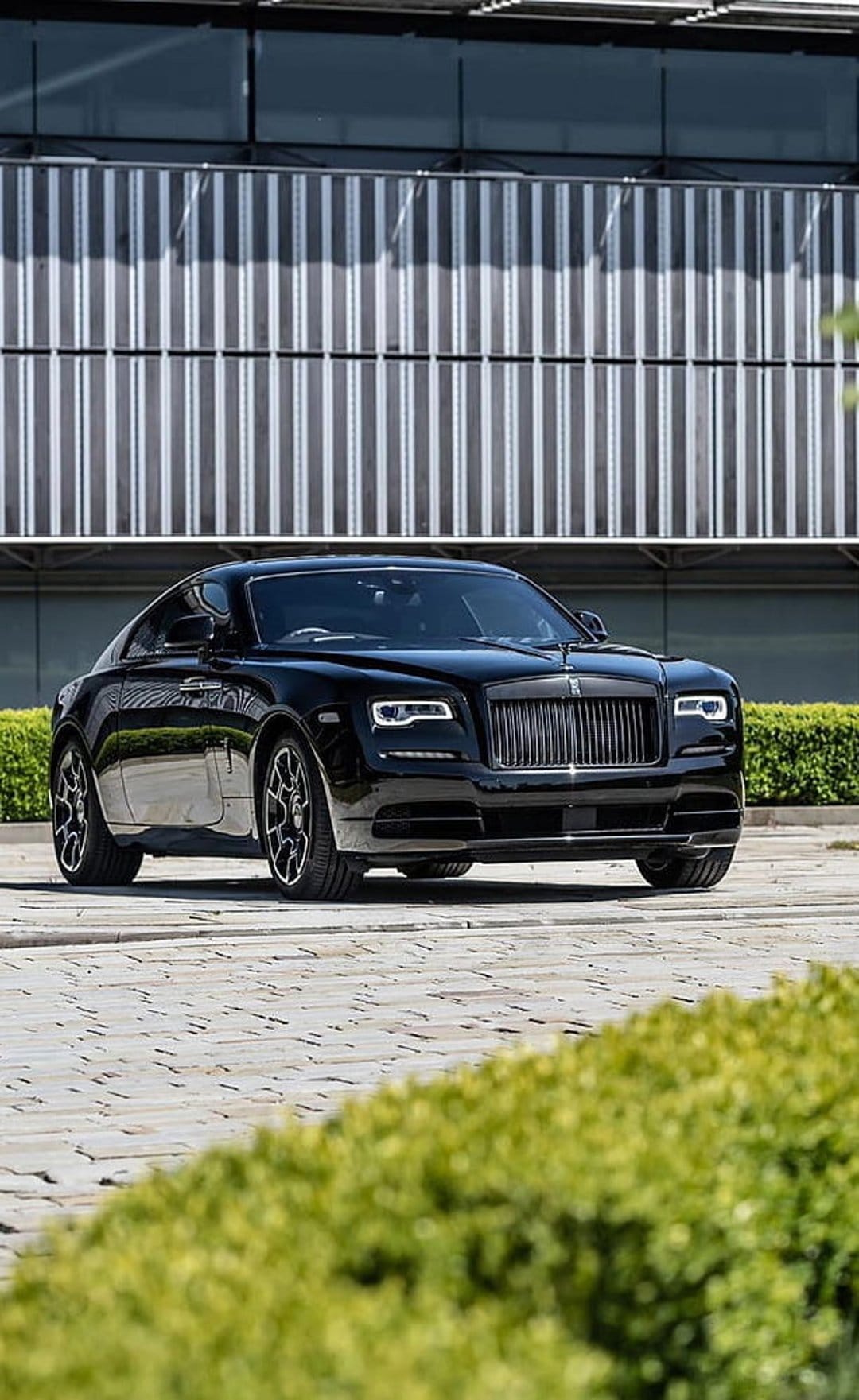 Black Rolls-Royce Wraith parked on a paved driveway in front of a modern building with silver vertical panels.