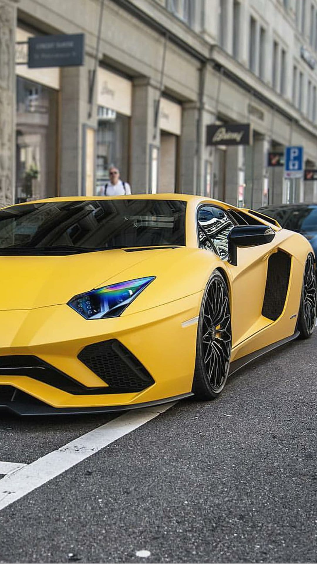 Yellow Lamborghini Aventador with LED headlights parked on a city street near buildings and a pedestrian.