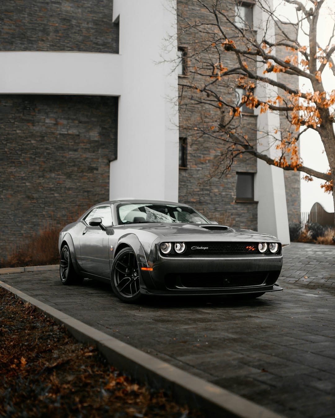 Grey Dodge Challenger Widebody parked on a concrete driveway near a modern brick building and autumn tree.