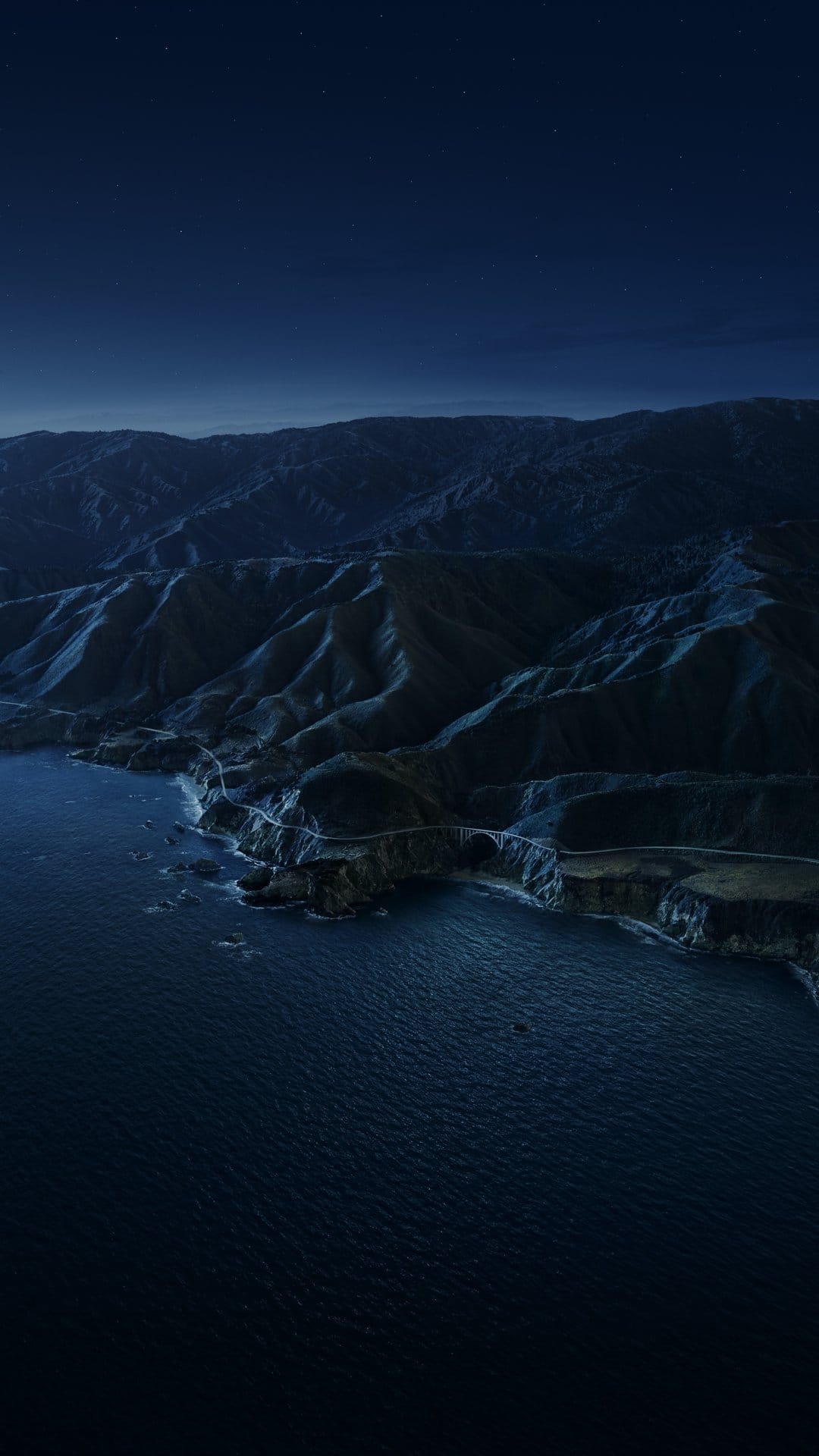 Winding coastal road snakes along steep cliffs beside the dark Pacific Ocean under a deep blue twilight sky.