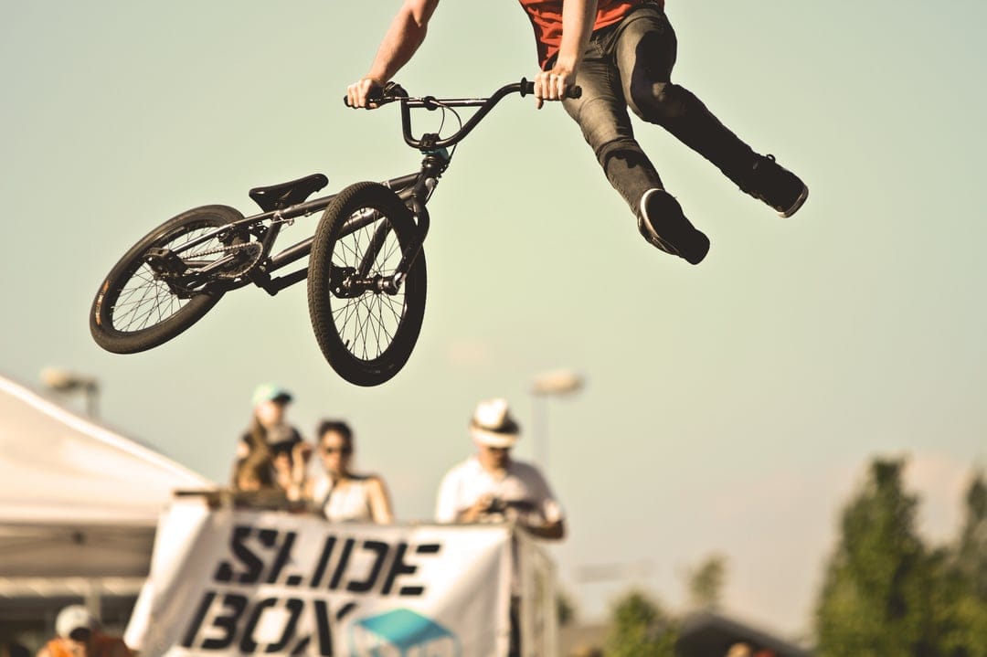 BMX rider performing a mid-air bicycle trick above a large crowd at an outdoor extreme sports competition.
