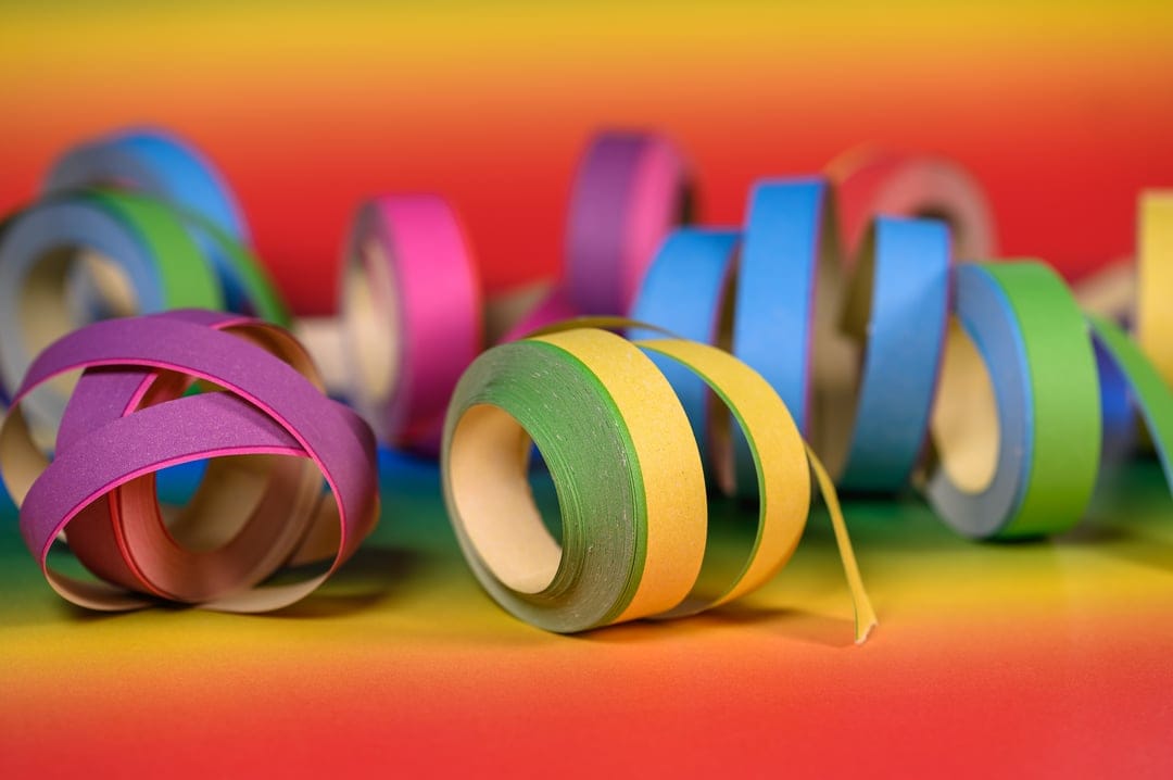 Colorful coiled streamers and paper ribbons over a soft-focus rainbow background with bright festive lighting.