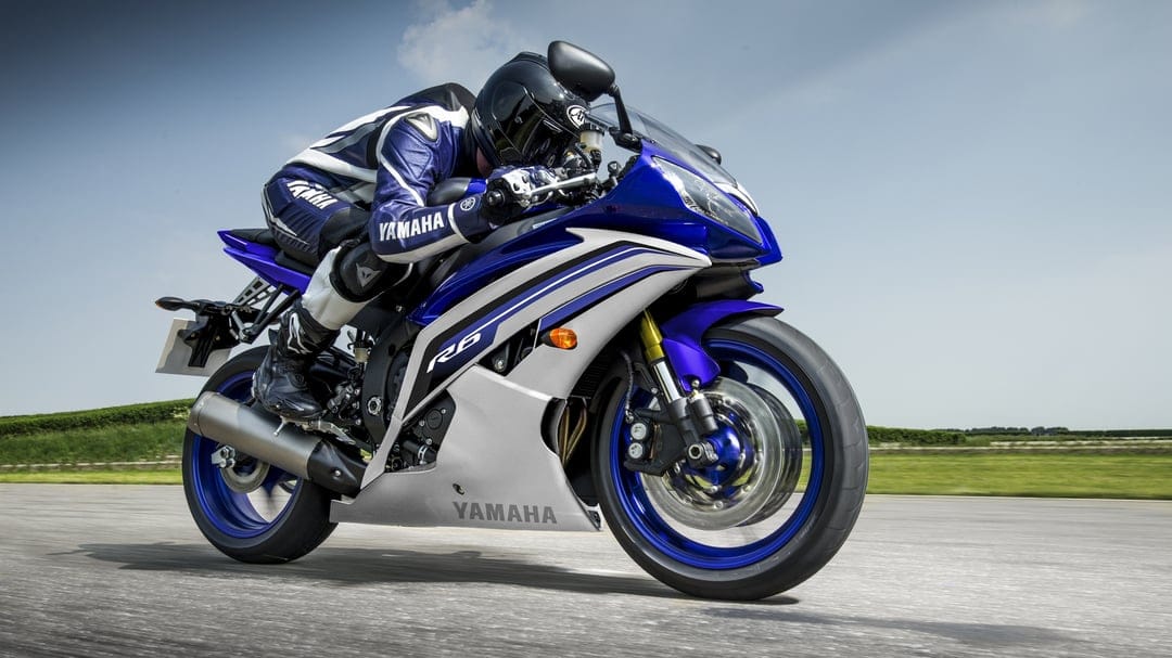 Blue Yamaha R6 motorcycle leaning into a turn with a rider in blue racing leathers on a paved track under sunlight.